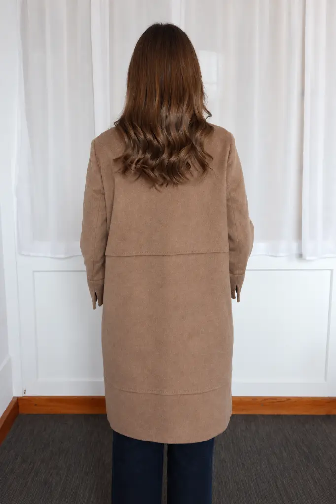 Manteau camel devant des rideaux blancs.webp